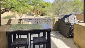 Une terrasse avec une table et des chaises, un barbecue abrité et une vue sur un paysage désertique, avec des bâtiments en arrière-plan.