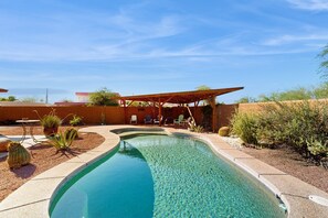 Piscina com borda curva e águas cristalinas, rodeada por um jardim bem cuidado com diversas plantas e uma área de estar com cadeiras.