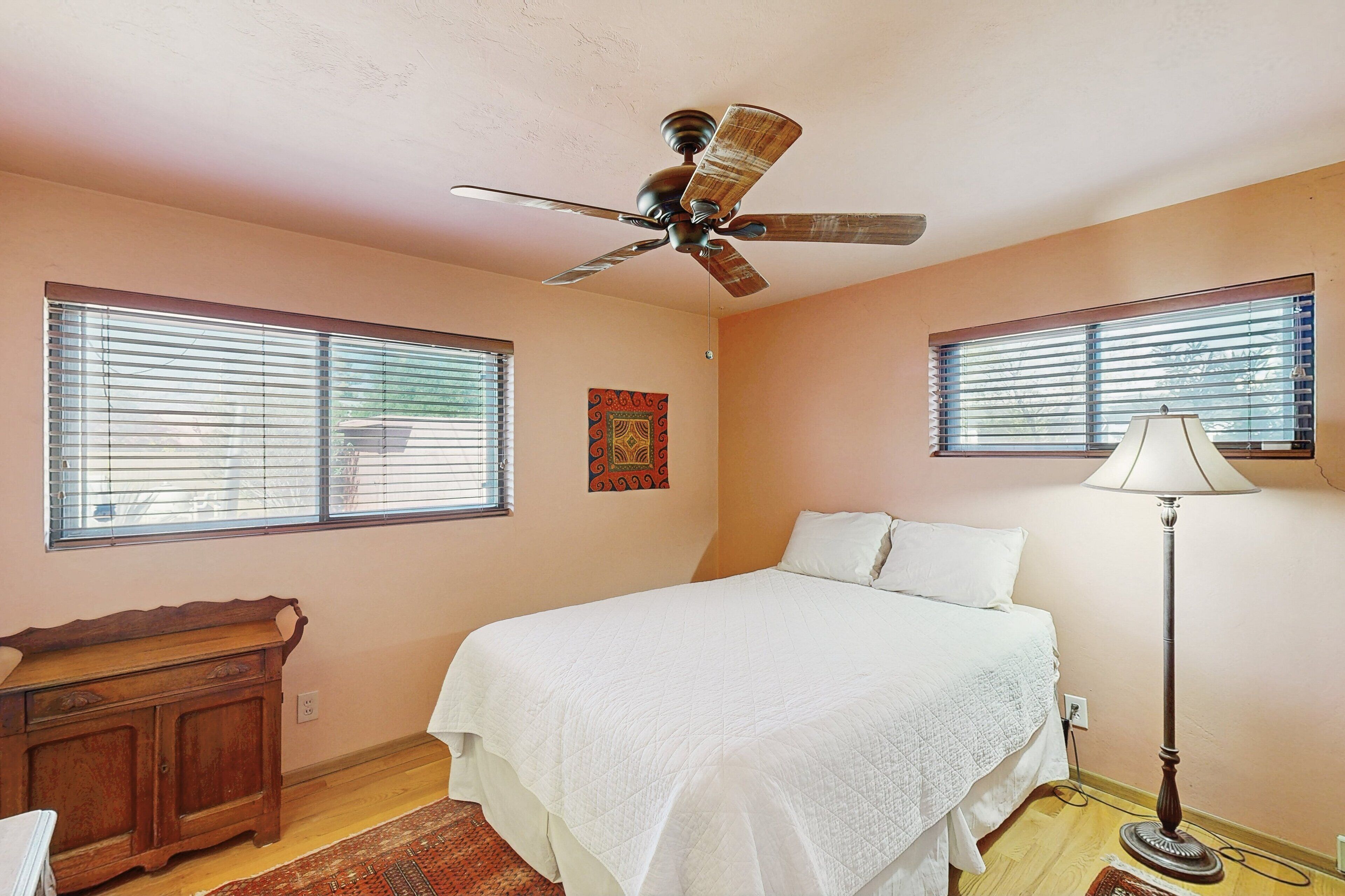 A bedroom with a bed, a ceiling fan, a nightstand, a lamp, and two windows with blinds.