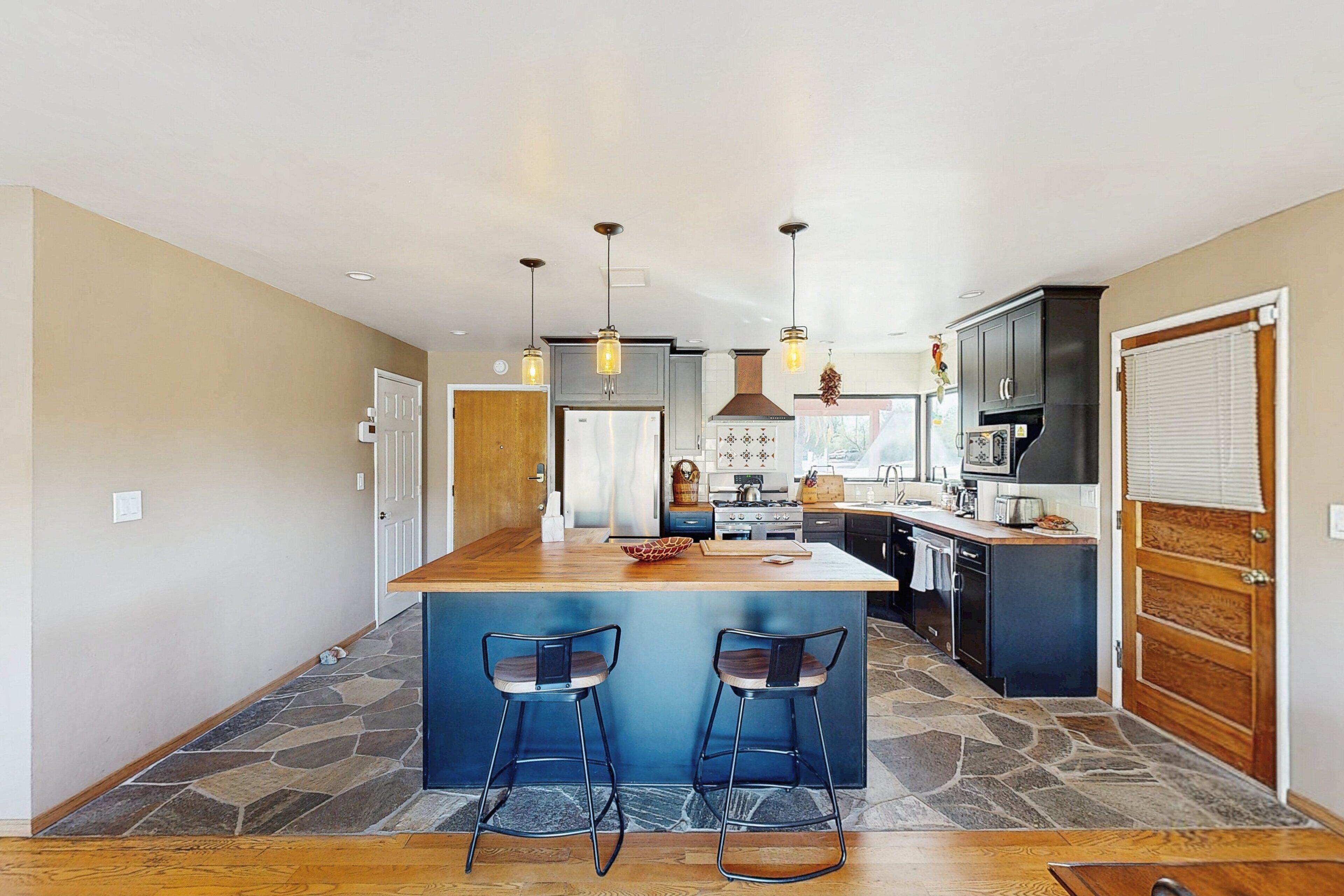 A modern kitchen with a central island, bar stools, pendant lighting, and a stone floor.