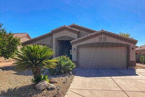 A residential house with a garage, a driveway, and landscaping including a palm tree and rocks. - Spacious desert home with hot tub, covered patio, grill, & modern finishes (Surprise)