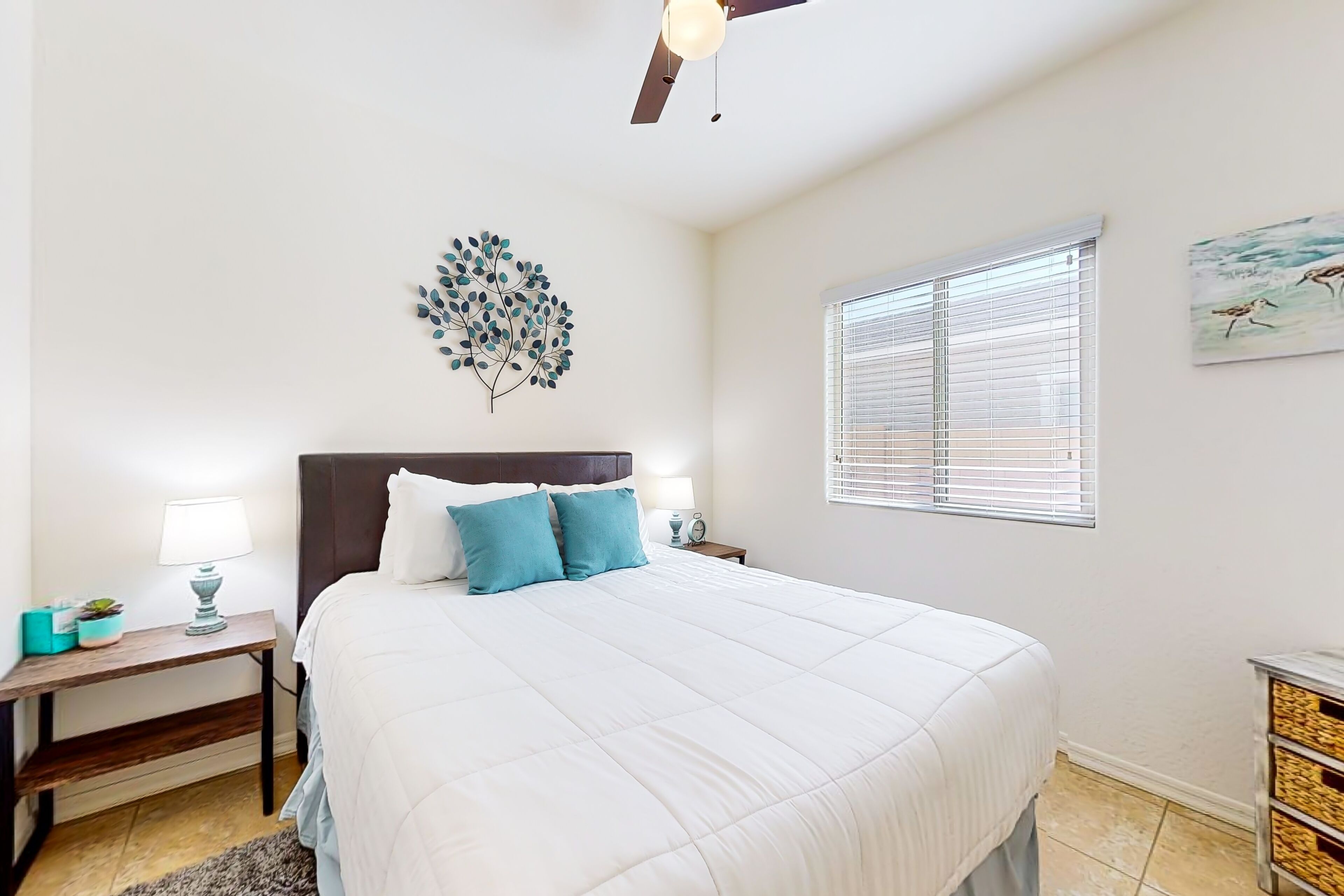 A bedroom with a bed, bedside tables, a wall art piece, a window with blinds, and a ceiling fan.