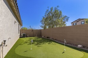 A well-maintained artificial green lawn with two golf flags, surrounded by a brick wall and a tree. - Airy desert home with an enclosed yard, private pool, hot tub, firepit, & W/D (Buckeye)