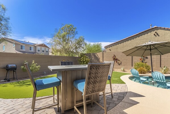 An outdoor patio with a bar, chairs, and a pool area. - Airy desert home with an enclosed yard, private pool, hot tub, firepit, & W/D (Buckeye)