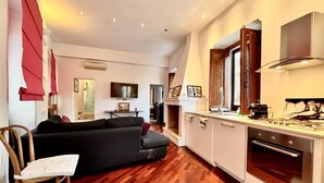 Uma sala de estar moderna com sofá preto, piso de madeira, lareira e cozinha com armários brancos e fogão.