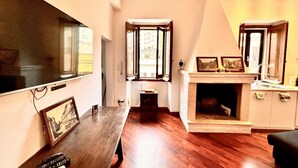Quarto com piso de madeira, lareira, televisão e janela com vista para edifícios.