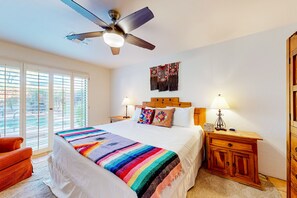 A bedroom with a large bed, a ceiling fan, a wooden nightstand with a lamp, and a view through the window.