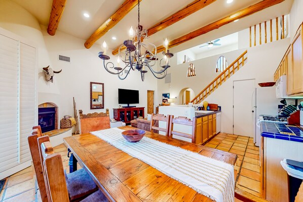 A spacious living room with a wooden dining table, a chandelier, and a kitchen in the background.