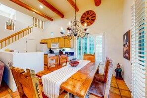 A spacious dining area with a wooden table, chairs, and a chandelier. A kitchen is visible in the background with wooden cabinets and a refrigerator.