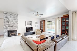 A modern living room with a stone fireplace, a large leather sofa, a coffee table, a TV, and a bookshelf. - Recently Remodeled Home | Fenced Backyard, Heated Pool, Grill & Fire Pit (Palm Springs)