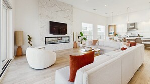 A modern living room with a sectional sofa, a round coffee table, and a flat-screen TV mounted on a marble wall. - Desert days, cozy nights, and poolside delights await at this stylish home. (Palm Springs)