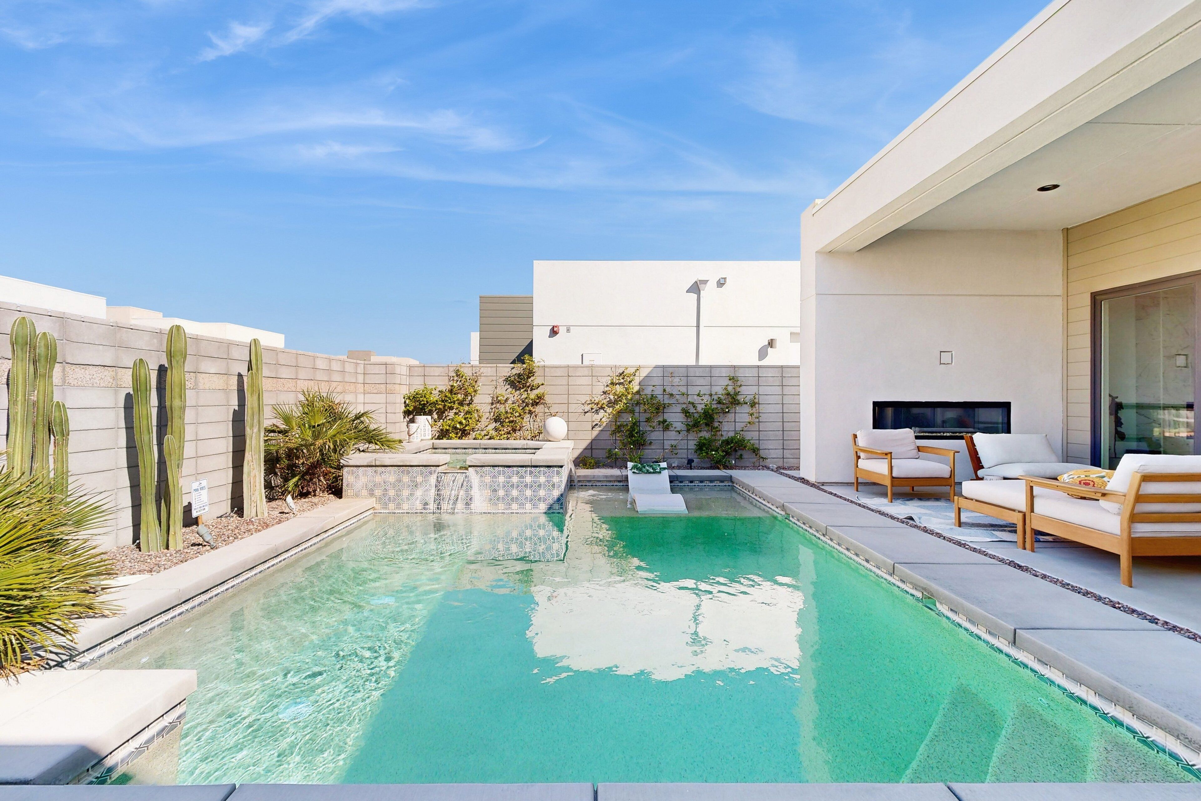 A modern outdoor pool area with a clear blue pool, surrounded by a low wall with cacti and small plants. There are two wooden lounge chairs and a small table with cushions.