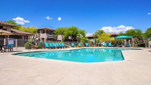 Ein Swimmingpool mit blauen Stühlen und Sonnenschirmen, umgeben von Gebäuden mit Ziegeldächern und viel Grün.