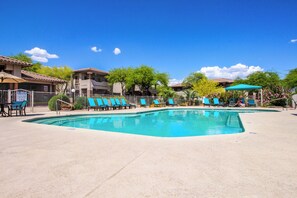 A swimming pool with blue chairs and umbrellas, surrounded by buildings with tiled roofs and greenery. - Tastefully remodeled condo with pool, hot tub & fitness room (Oro Valley)