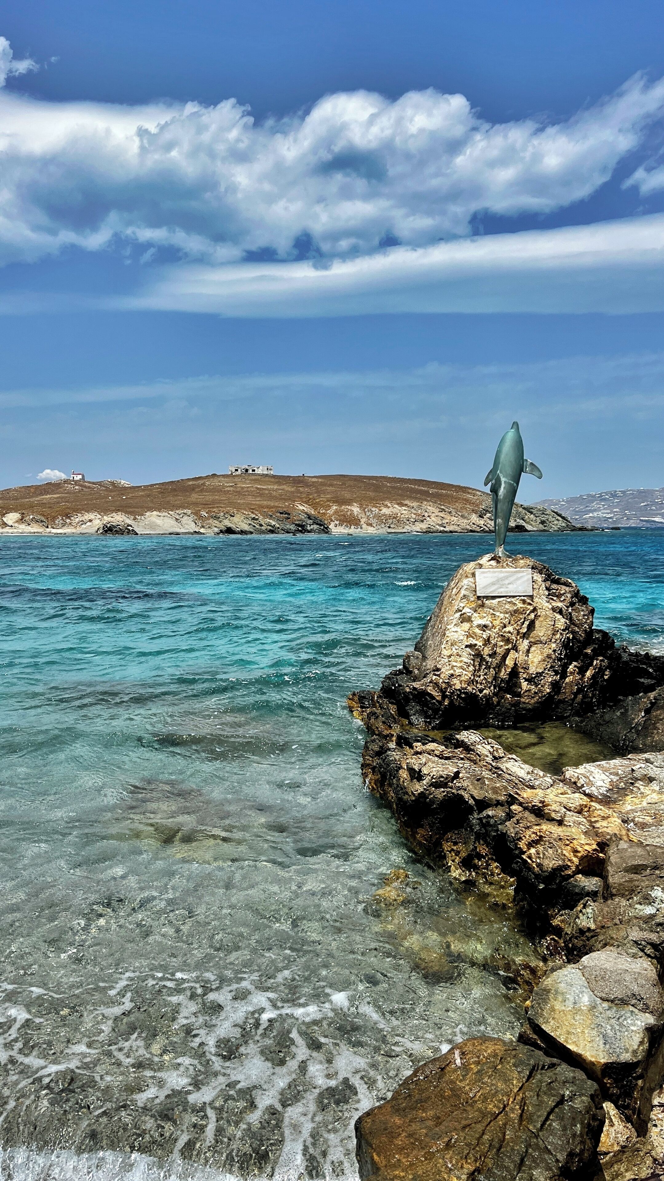 Un rivage rocheux bordé d’une eau claire, une statue et une colline qui se dessine à l’horizon.