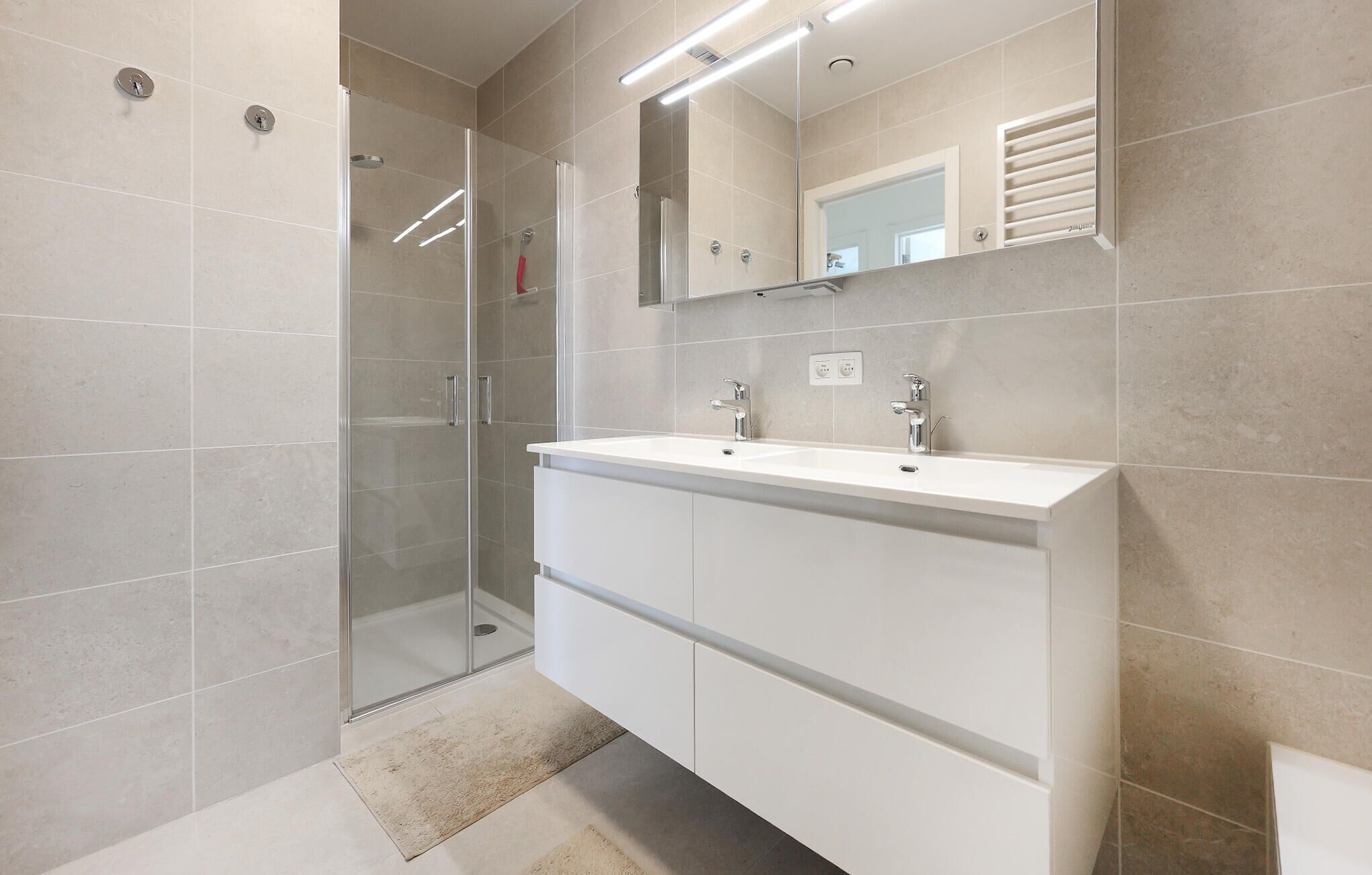 Een moderne badkamer met een douchecabine van glas, een wit wastafelmeubel met twee wastafels en een grote spiegel erboven.