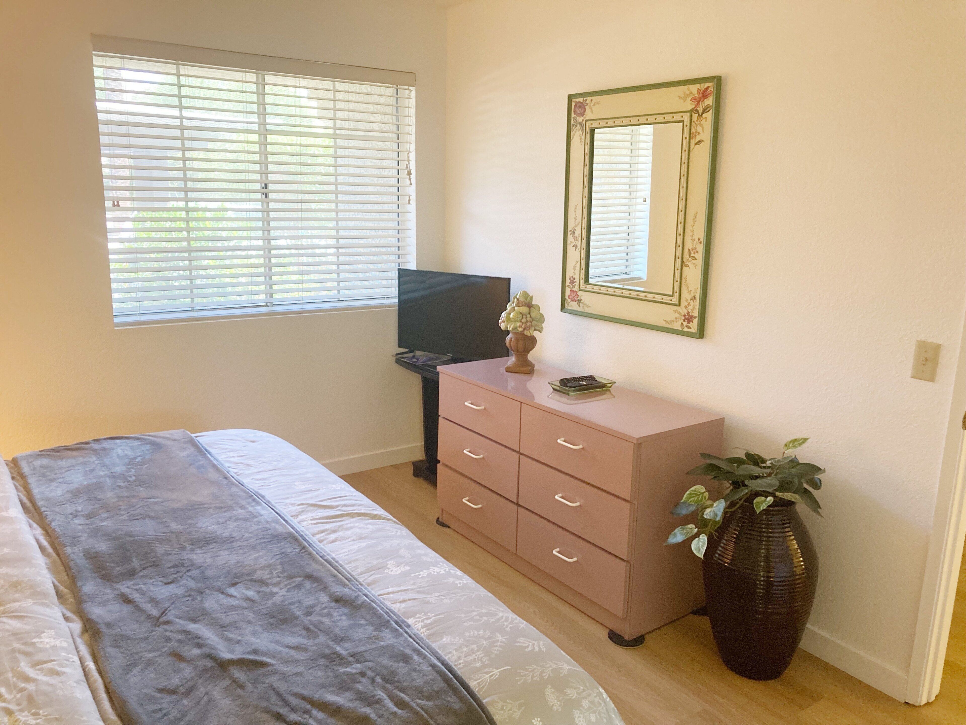 A bedroom with a bed, dresser, TV, and a window with blinds.