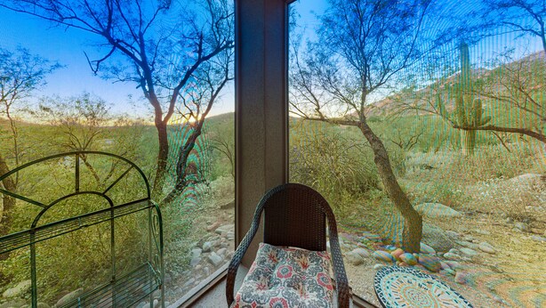 Ein Zimmer mit einem Korbstuhl und einem Tisch mit Muster, ein Fenster mit Blick auf Bäume und eine Wüstenlandschaft.