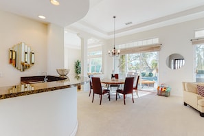 Cozinha moderna com ilha curva, área de jantar com mesa e cadeiras redondas, e sala de estar com sofá e janela grande.