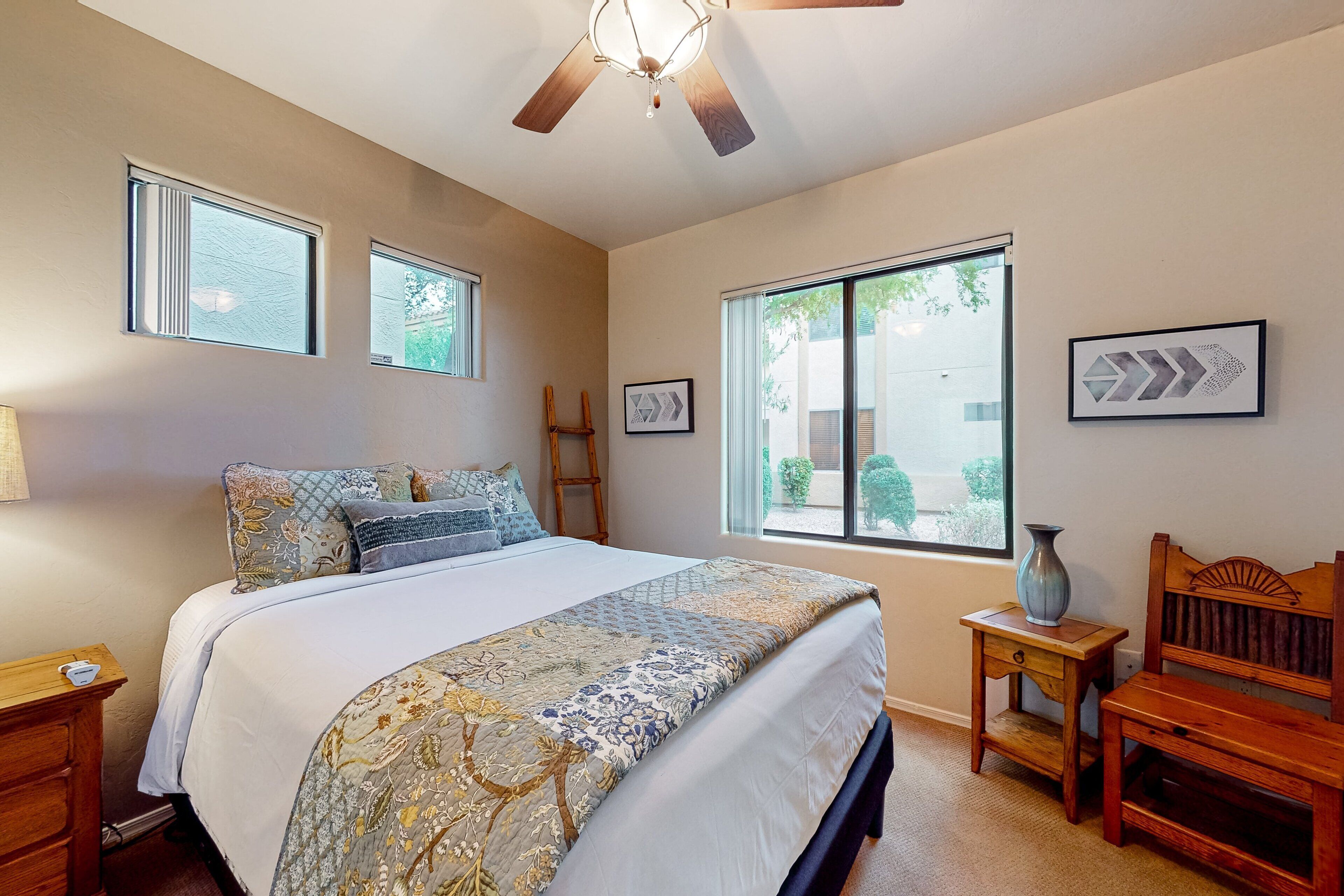 A bedroom with a bed, two windows, a ceiling fan, a nightstand, a bedside table, a lamp, and a chair.