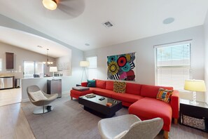 A modern living room with a red sofa, a glass coffee table, and a dining area with a kitchen in the background. - Pristine Desert Ridge home with a pool & backyard oasis - dog-friendly (Phoenix)