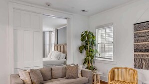 A living room with a sofa, a wicker chair, a small table, and a large plant. - Historic, Spacious Loft w. Kitchen + Living Room (Charleston)