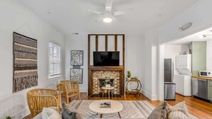 A modern living room with a fireplace, a television, wicker furniture, and a kitchen in the background. - Historic, Spacious Loft w. Kitchen + Living Room (Charleston)