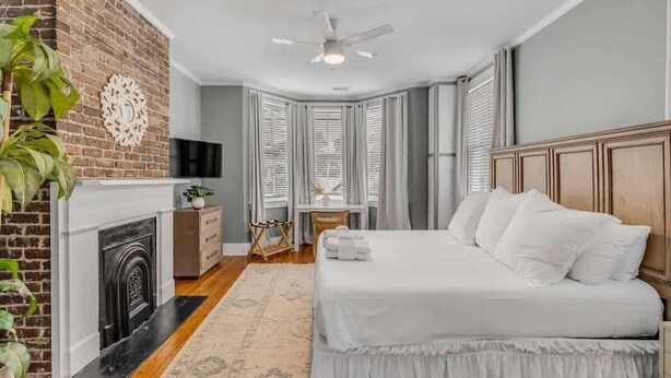 A bedroom with a brick fireplace, a bed with white bedding, a ceiling fan, and a view of the city through large windows. - Historic, Spacious Loft w. Kitchen + Living Room (Charleston)