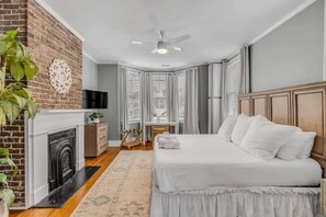 A bedroom with a brick fireplace, a bed with white bedding, a ceiling fan, and a view of the city through large windows. - Historic, Spacious Loft w. Kitchen + Living Room (Charleston)