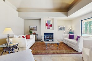 A modern living room with white sofas, a glass coffee table, a fireplace, and a large window with a view of trees. - Desert Bliss with Mountain Views & a Sparkling Private Pool. (Palm Desert)