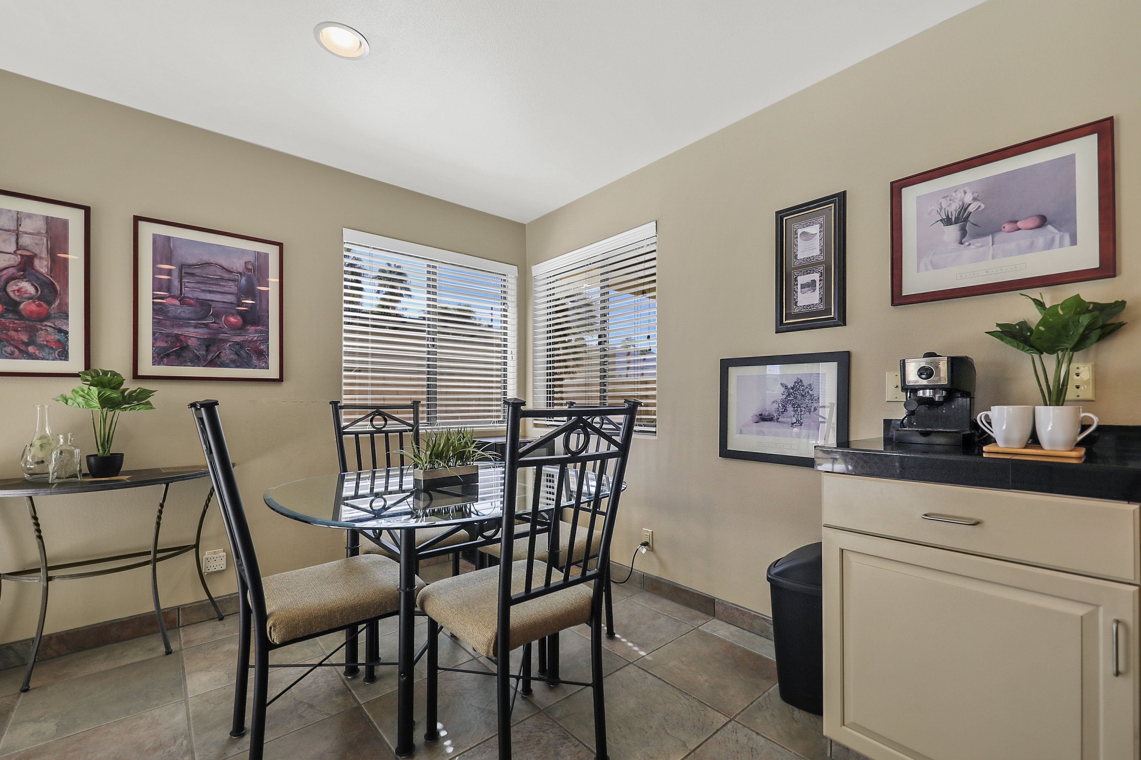 A dining area with a glass table, black chairs, and a black countertop with a coffee machine. There are framed pictures on the wall and a potted plant on the countertop.