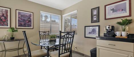 A dining area with a glass table, black chairs, and a black countertop with a coffee machine. There are framed pictures on the wall and a potted plant on the countertop.