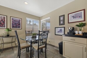 A dining area with a glass table, black chairs, and a black countertop with a coffee machine. There are framed pictures on the wall and a potted plant on the countertop. - Desert Bliss with Mountain Views & a Sparkling Private Pool. (Palm Desert)