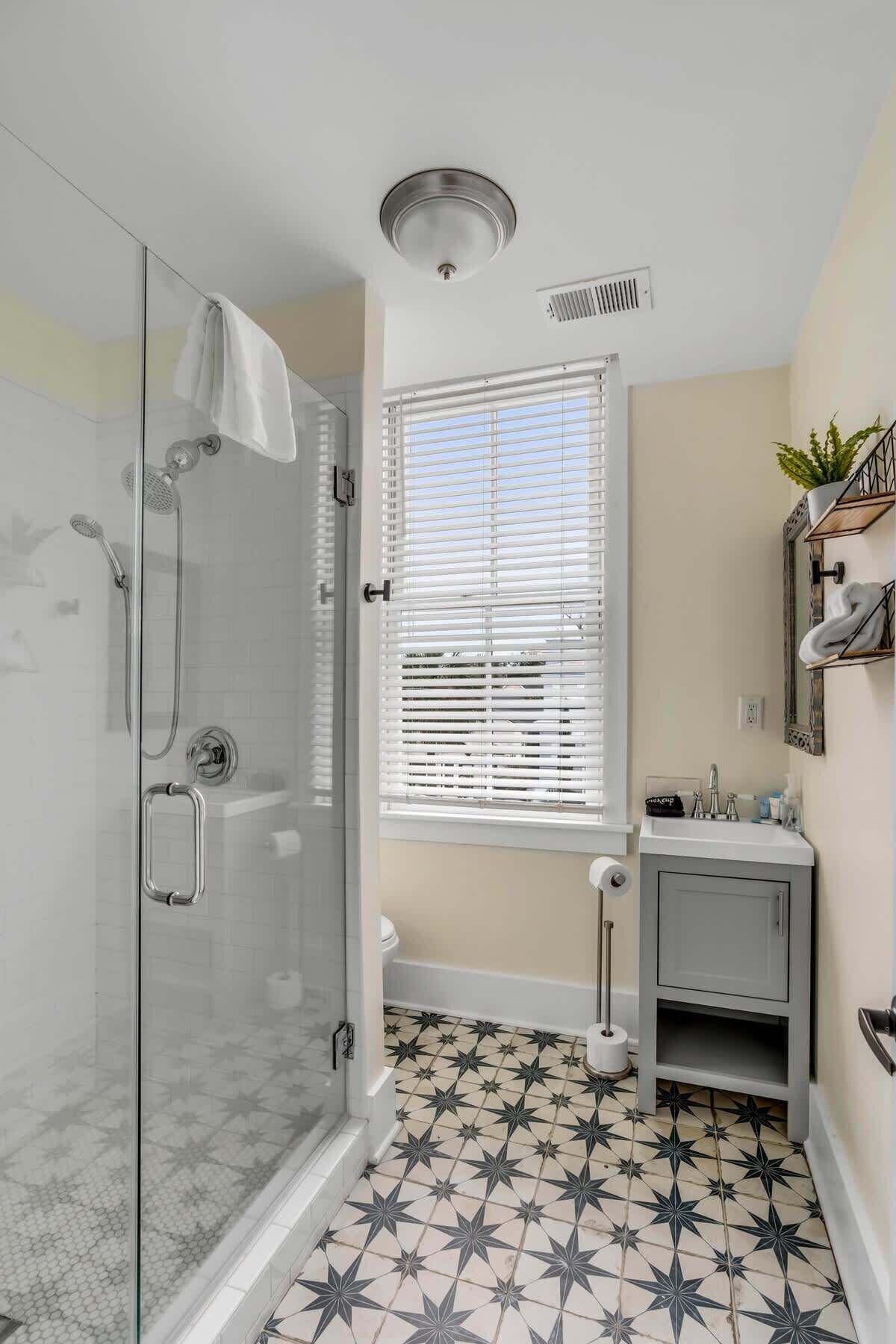 Une salle de bain avec une douche à parois de verre, des toilettes, un lavabo avec un miroir et une fenêtre avec des stores.