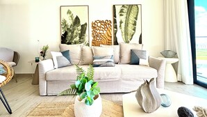 Een moderne woonkamer met een beige bank, decoratieve kussens, een grote plant en kunst aan de muur.
