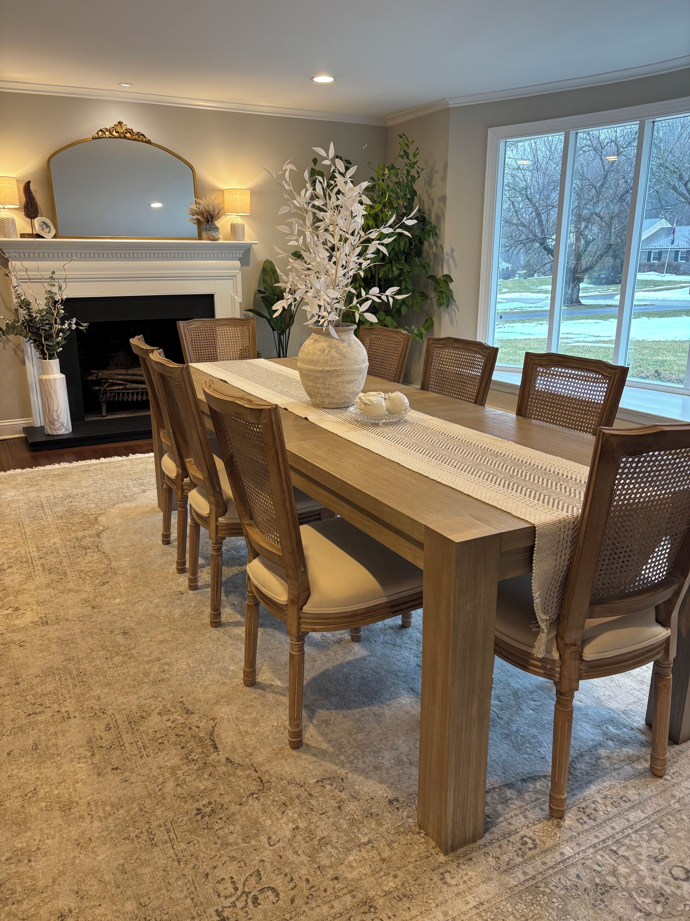 Uma sala de jantar com mesa de madeira, seis cadeiras, um vaso grande com flores brancas, lareira e uma janela ampla com vista para o exterior.