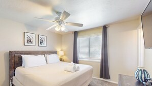 Ein Hotelzimmer mit einem großen Bett, einem Deckenventilator, zwei gerahmten Kunstwerken an der Wand, einem Fenster mit Jalousien und einem kleinen Tisch mit einer Vase.