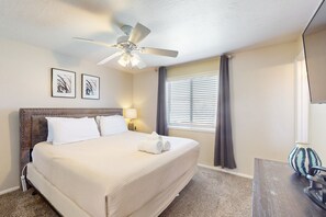 Ein Hotelzimmer mit einem großen Bett, einem Deckenventilator, zwei gerahmten Kunstwerken an der Wand, einem Fenster mit Jalousien und einem kleinen Tisch mit einer Vase.