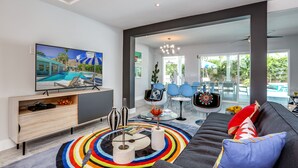 Un salon moderne avec une grande télévision, un canapé, un tapis coloré et un coin repas avec des chaises bleues.