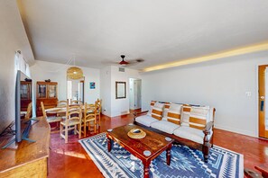 A living room with a wooden coffee table, a sofa with striped cushions, a dining area with wooden chairs, and a ceiling fan. - Charming & bright bungalow w/ patio, yard, close to downtown shops, dogs ok (Tucson)