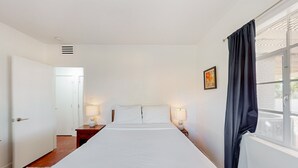Ett hotellrum med en stor säng, två sänglampor, ett fönster med gardiner och en dörr som leder till ett annat rum.