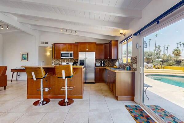 A modern kitchen with wooden cabinets, stainless steel appliances, and a bar area with stools. - Contemporary house with pool, spa, firepit, gas grill, W/D, & central AC (Rancho Mirage)