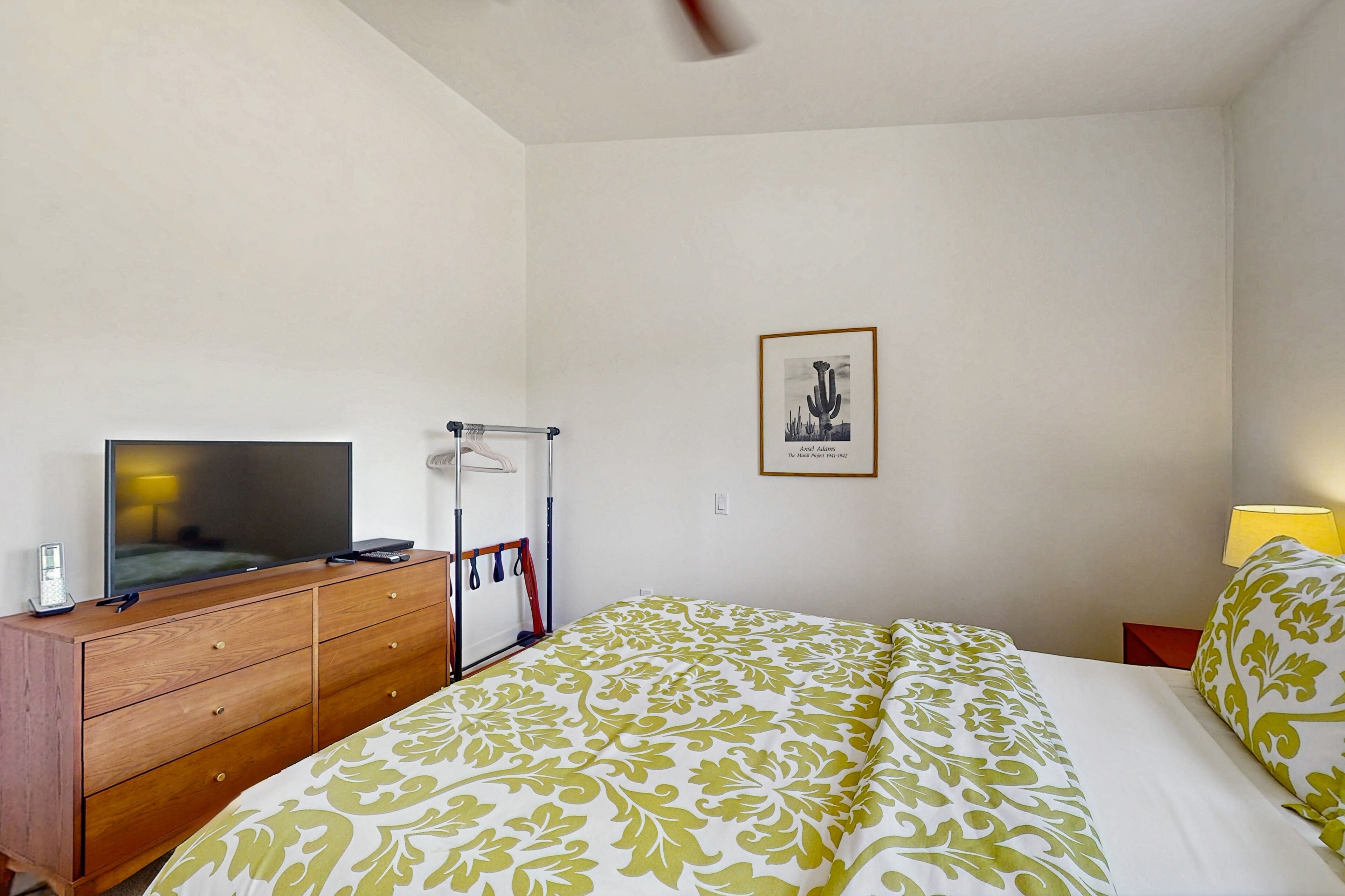 A bedroom with a bed, a wooden dresser, a television, a framed picture on the wall, and a lamp on the nightstand.