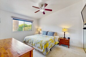 Une chambre à coucher avec un lit, deux tables de chevet avec des lampes, un ventilateur de plafond, une fenêtre donnant sur un beau paysage et un bureau en bois.