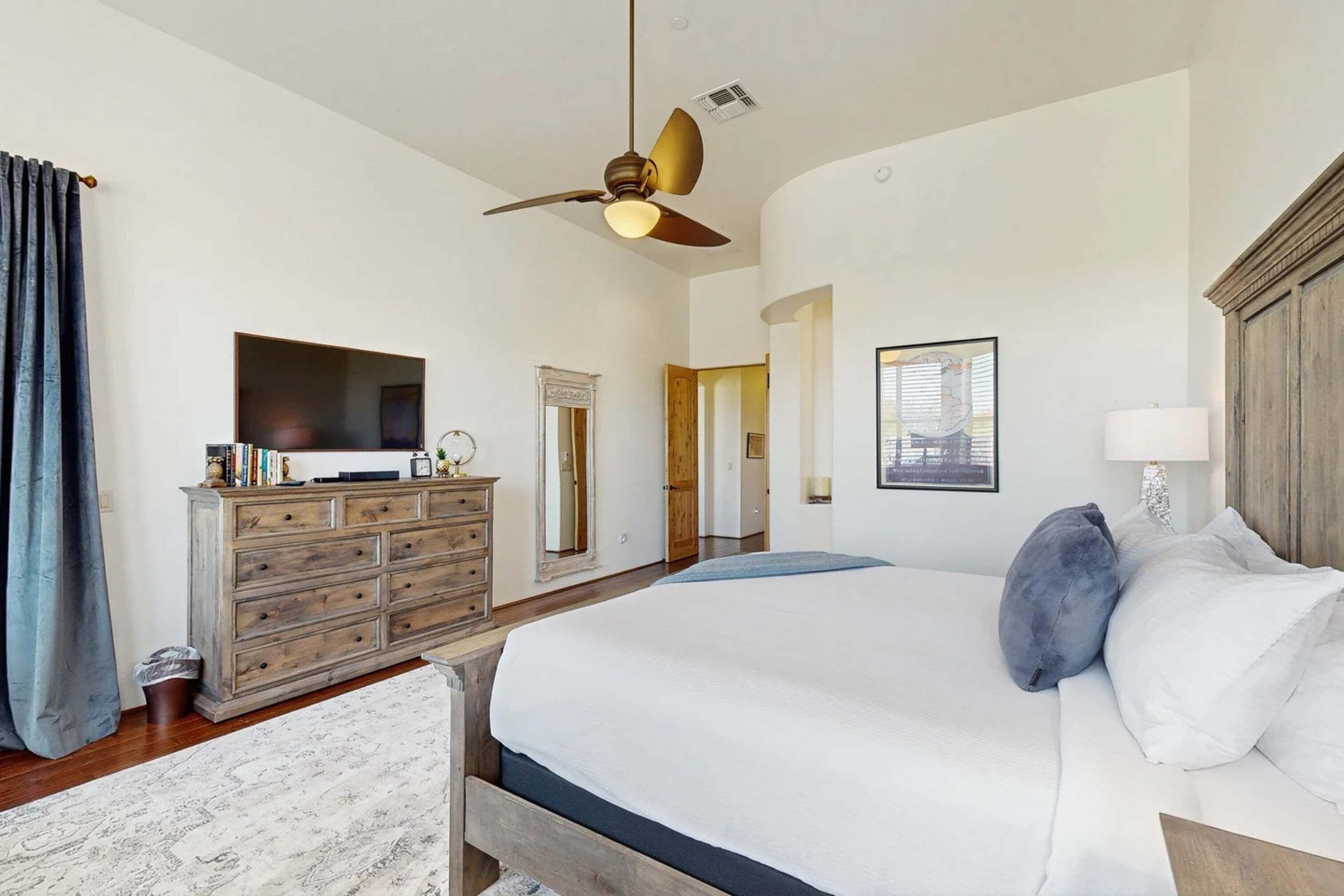 A bedroom with a large bed, a ceiling fan, a dresser, a television, and a door leading to another room.