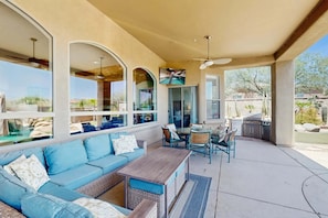 An outdoor patio with a blue sofa, wicker furniture, a wooden table, and a TV mounted on the wall. - Dog-Friendly Desert Oasis w/ a Gourmet Kitchen, Private Pool, & Putting Green (Marana)