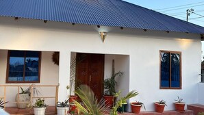 A single-story house with a corrugated metal roof, a front door, and a small porch area with potted plants.