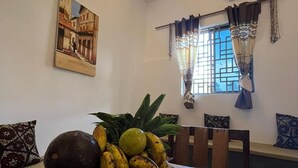 A dining area with a wooden table, a fruit bowl, a wall-mounted air conditioner, patterned curtains, and a painting.