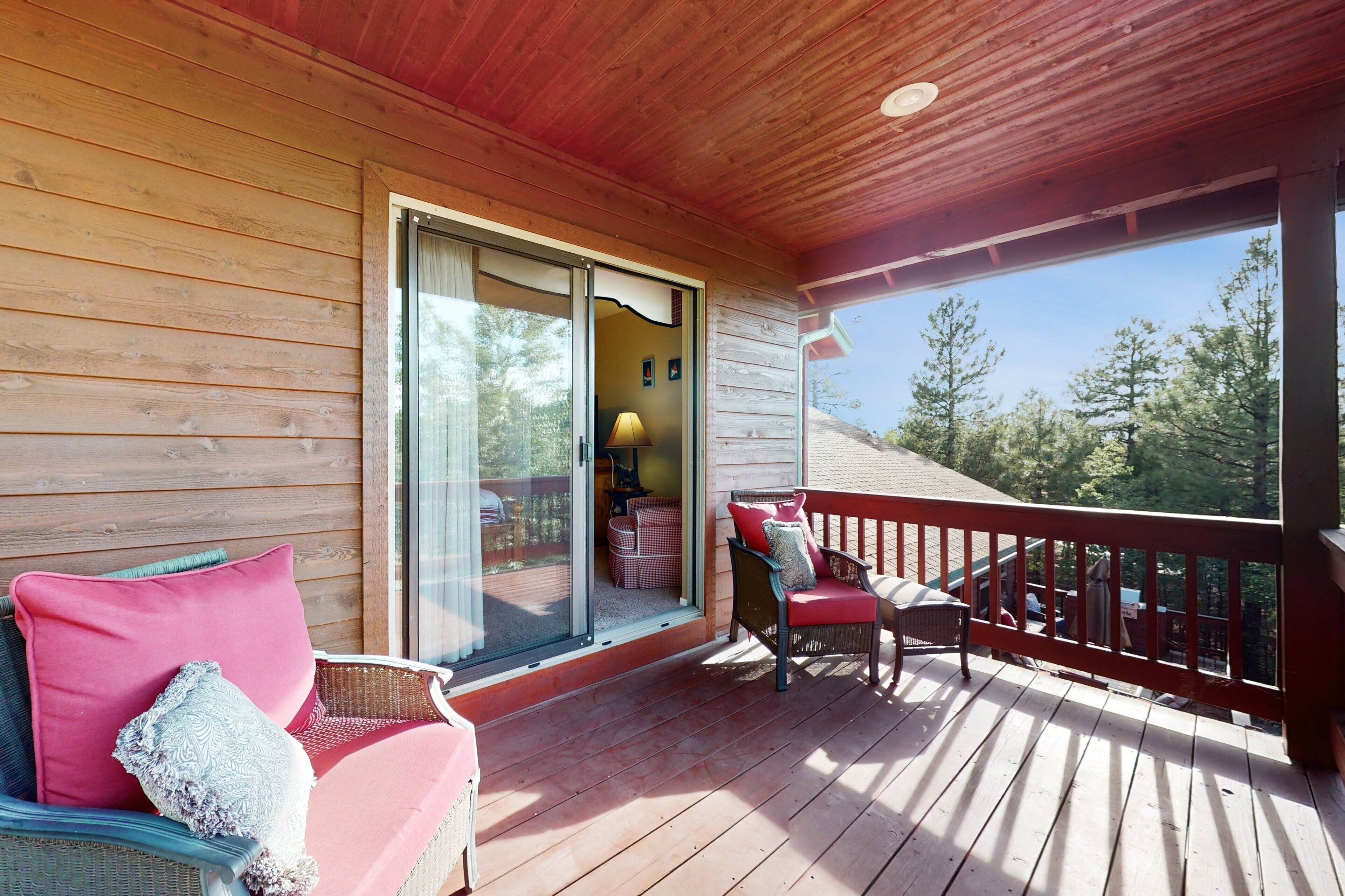 A wooden deck with a covered area, a wicker chair with a red cushion, and a view of trees through a glass door.