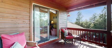 A wooden deck with a covered area, a wicker chair with a red cushion, and a view of trees through a glass door.